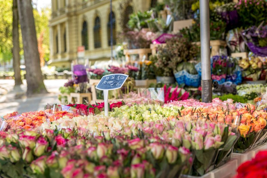 A vibrant outdoor flower market with an array of fresh floral bouquets in pink, peach, and white roses, displayed on wooden tables. The flowers are tightly packed, showcasing their delicate petals and