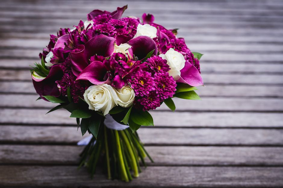 A bouquet of fresh, vibrant flowers arranged on a wooden surface, featuring large deep purple calla lilies, small white roses, and clusters of bright magenta chrysanthemums and hydrangeas. The bouquet