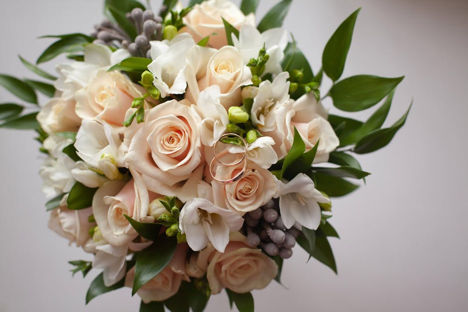 A close-up of a floral bouquet arranged with pale peach roses, white freesias, and small clusters of grey berries, complemented by an abundance of green foliage, set against a neutral background. The