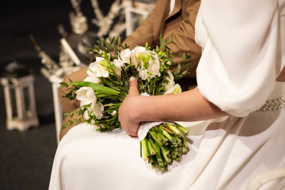 A bride in a white wedding dress and a person in a brown outfit holding a bouquet of fresh white eustoma flowers with green foliage. The bouquet is wrapped with white lace at the stems, and the scene