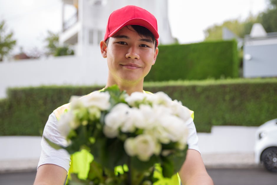 A delivery person wearing a bright red cap and a high-visibility yellow vest is smiling as he offers a bouquet of fresh white flowers, including roses and greenery, wrapped in clear cellophane, toward