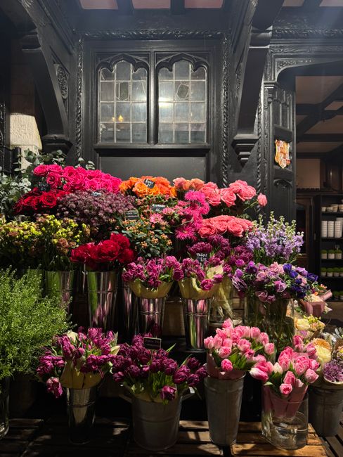 An arrangement of fresh flowers displayed on a dark wooden shelf inside a florist shop. The bouquet includes vibrant pink, purple, red, and orange blooms, featuring carnations, chrysanthemums, and oth