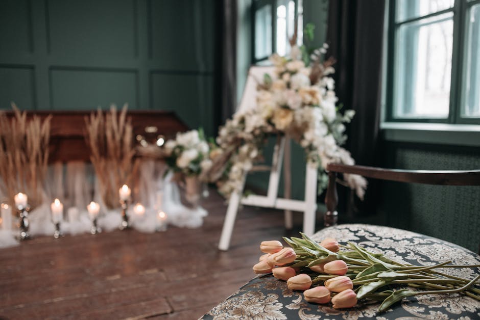 A floral arrangement for a funeral setting, featuring white and cream-colored flowers such as roses and lilies, placed on a wooden floor with a dark green paneled wall background. Soft natural light f