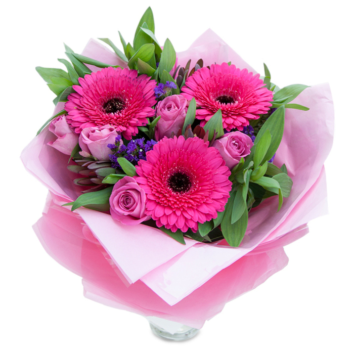 I'd describe this composition as playfulness made floral, the kind of bouquet that reflects both charm and considered craftsmanship you'd expect from a local Castelnau florist. At the heart are three wide-eyed gerberas in a vivid fuchsia, their sculpted petals fanning out from deep dark centres that ground the arrangement with a striking contrast. Surrounding them, lavender-tinged roses in a dusty pink hold their petals close, creating soft, tactile spirals that invite a gentle touch. Scattered throughout are tiny clusters of purple statice, their fine, textured blooms offering sparks of cool colour and a meadowlike quality. Lush green foliage-broad leaves resembling salal or eucalyptus-wraps around the flowers, adding depth, a clean green scent, and a subtly dewy visual. The bouquet is thoughtfully wrapped: an inner sheet of soft matte pale pink tissue supports the blossoms, while an outer layer of translucent fuchsia-pink cellophane gives a slight sheen and a modern finish. The rim of a clear glass vase peeks out from the lower section, suggesting the arrangement is prepared for immediate display at a kitchen table or hallway stand. The lighting in the image is soft and natural, accentuating petal textures and the gentle interplay of warm and cool tones. Perfect for brightening a bedside, marking a birthday, or surprising a neighbour returning from a stroll along the riverside, this bouquet radiates approachable elegance and the kind of careful, garden-inspired composition that becomes a small, memorable celebration.